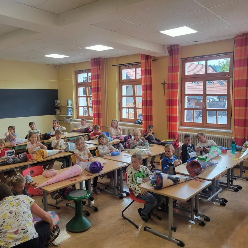 Ein Klassenzimmer voller kleiner Kinder, die an Schreibtischen sitzen, einige mit Gesichtsmasken, andere beschäftigen sich mit Aktivitäten. Der Raum hat gelbe Wände, große Fenster mit roten Vorhängen und einen Heizkörper.