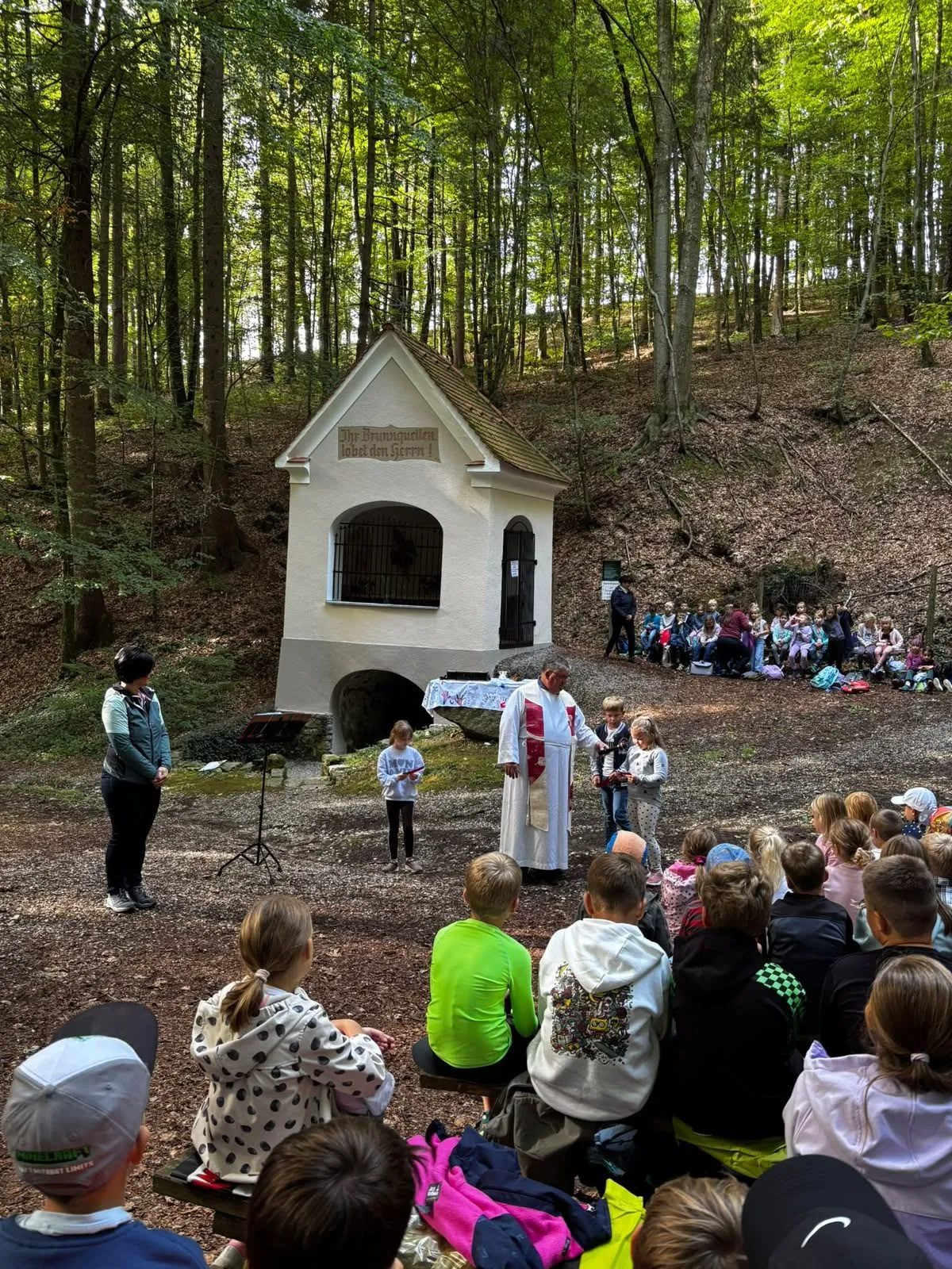 Ein Priester hält einen Gottesdienst in einer Waldkapelle ab, mit Kindern und Erwachsenen, die sich versammelt haben. Die Umgebung ist von Bäumen und einem kleinen Gebäude umgeben.