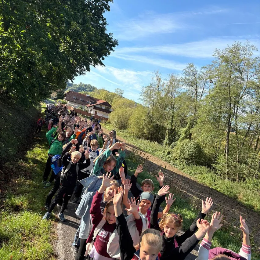 Eine große Gruppe von Kindern winkt an einem sonnigen Tag zur Kamera, während sie auf einem ländlichen Pfad stehen. Bäume und ein Haus sind im Hintergrund zu sehen.