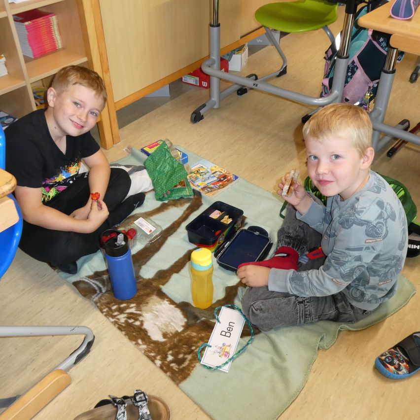 Zwei junge Jungen sitzen auf einem Teppich, einer mit einem Sandwich, der andere mit einem Becher. Sie haben Spielzeug und Bücher um sie herum auf einem Holzboden.
