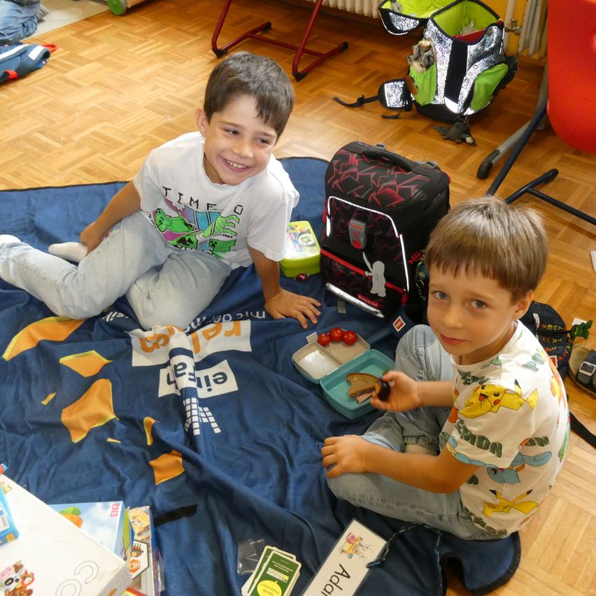 Zwei Jungen sitzen auf einer blauen Decke und lächeln. Einer hält ein Sandwich, der andere eine Brotdose. Spielzeug und ein Rucksack sind um sie herum.