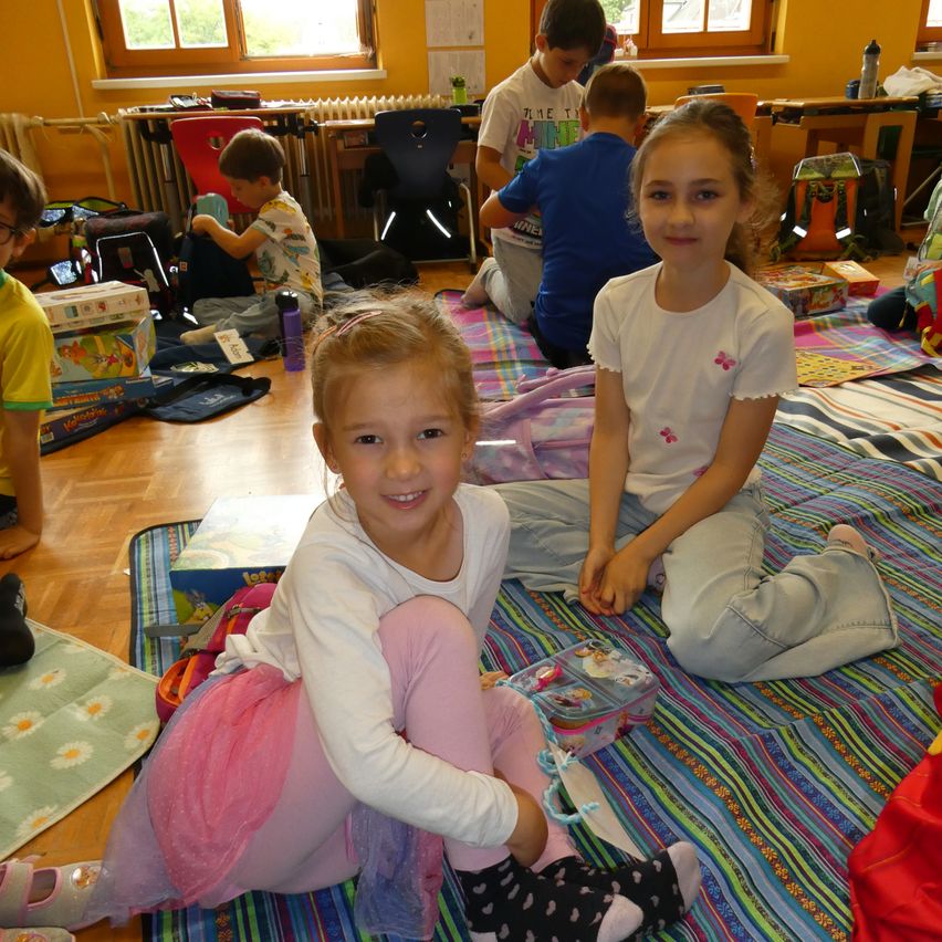 Kinder sitzen auf dem Boden in einem Klassenzimmer mit bunten Matten und Spielzeug um sie herum. Ein Mädchen lächelt und hält eine Tasche. Ein anderes Mädchen schaut zur Kamera.