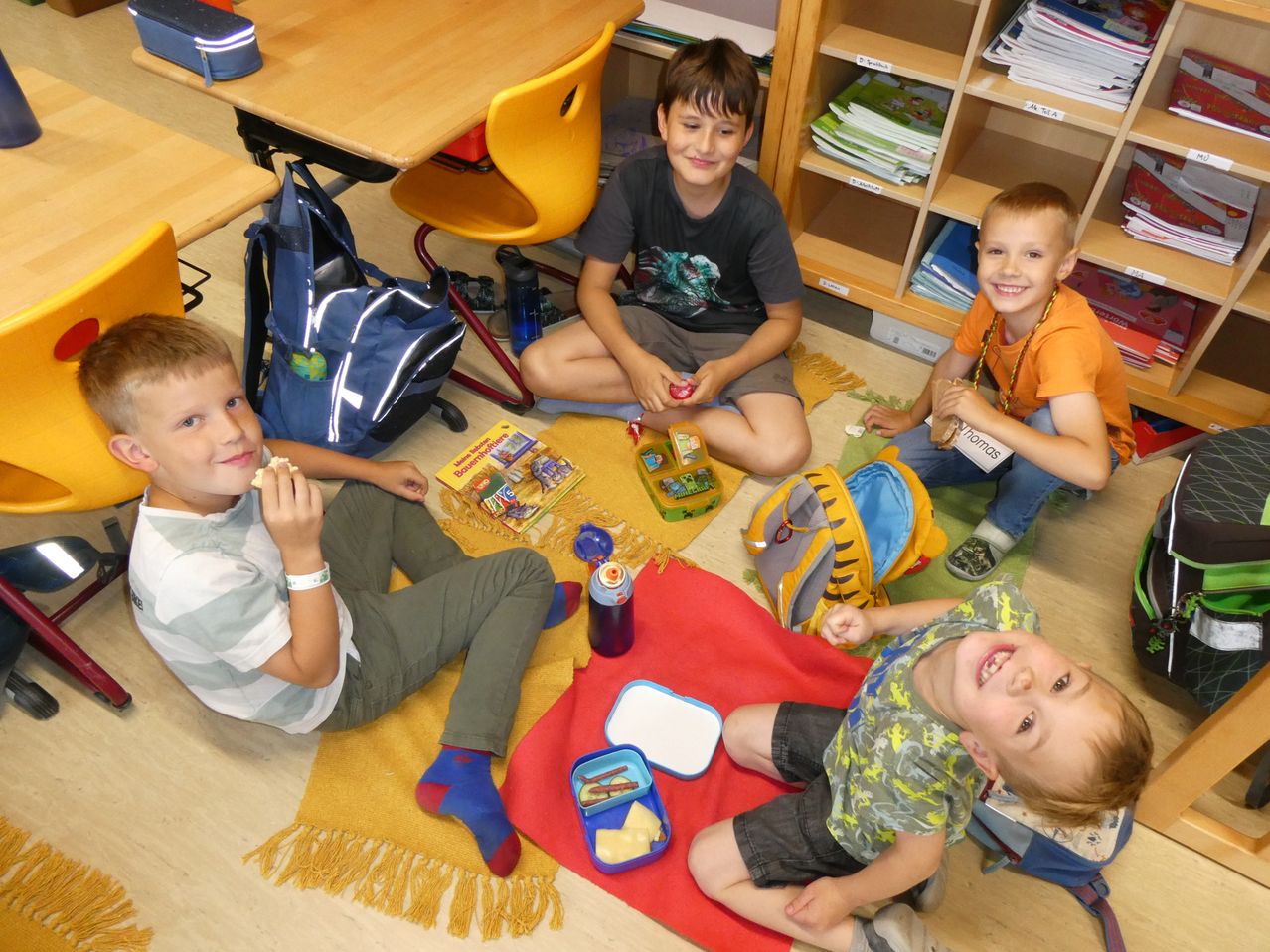 Vier junge Jungen sitzen auf einem Teppich und essen Snacks. Ein Junge hat einen blauen Rucksack. Ein anderer hat ein Buch. Ein dritter Junge trägt ein Armband.
