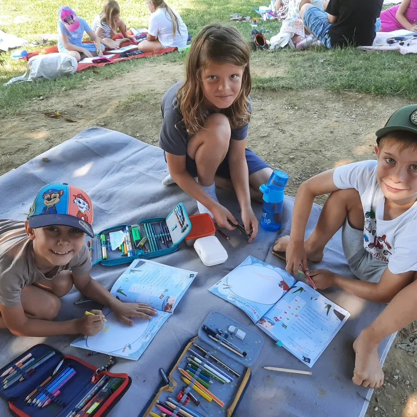 Drei Kinder sitzen auf einer Decke auf dem Rasen und machen Malaktivitäten. Sie haben Malbücher und Marker auf der Decke. Sie scheinen sich in einem Park zu befinden.