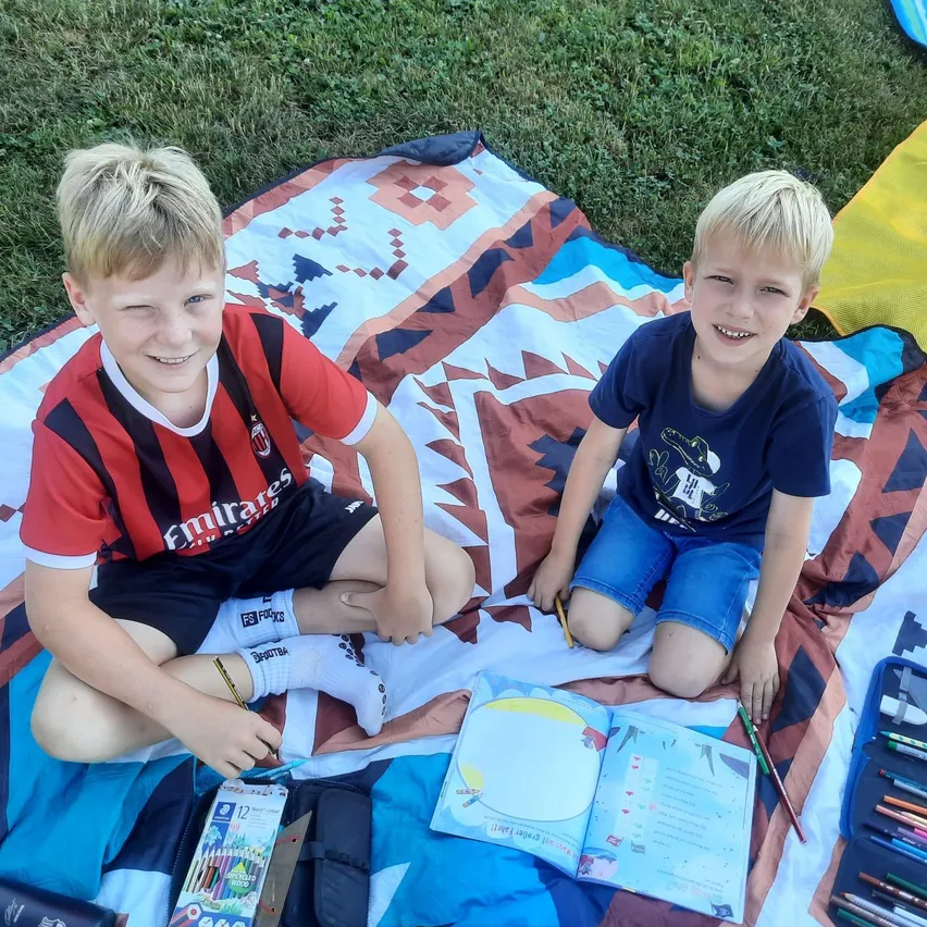 Zwei junge Jungen sitzen auf einer Decke mit verschiedenen Malutensilien und einem offenen Malbuch. Einer trägt ein rot-schwarz gestreiftes Shirt, der andere ein blaues.