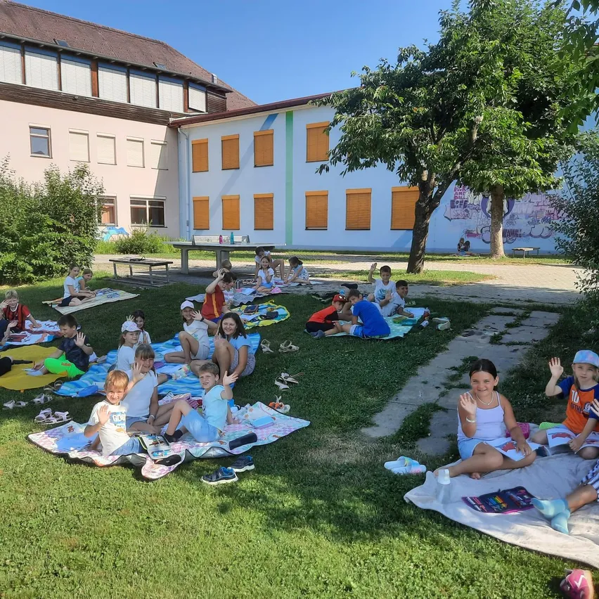 Kinder sitzen auf dem Rasen vor einem weißen Gebäude mit orangefarbenen Fensterläden, unter einem Baum, an einem sonnigen Tag.