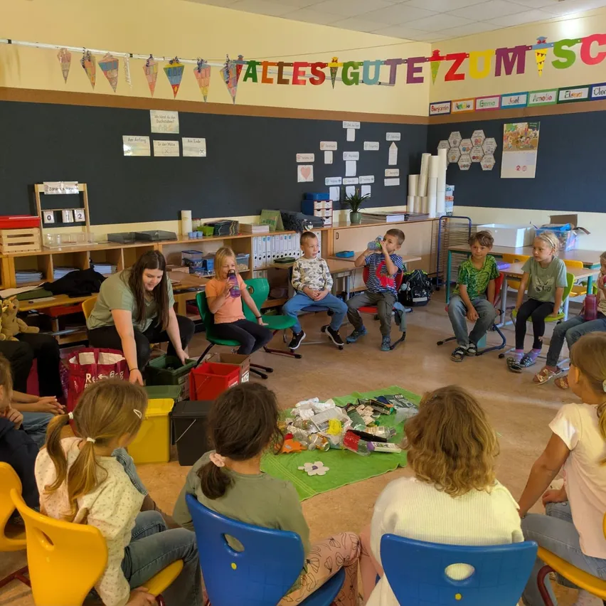 Ein Klassenzimmer mit Kindern, die auf Stühlen um einen Tisch herumsitzen, einige halten Tassen und andere schauen auf eine Frau, die vor ihnen steht.