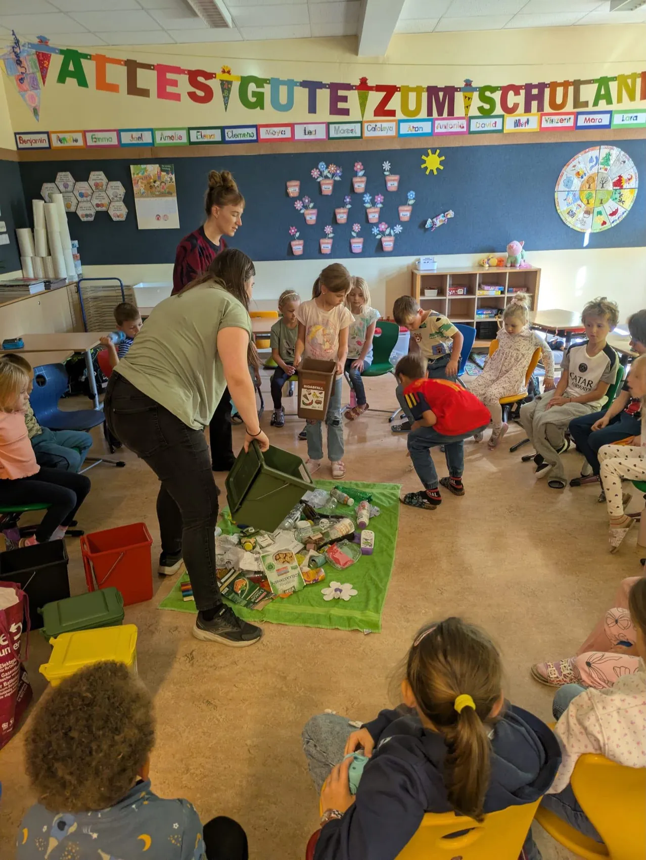 Eine Frau steht in einem Klassenzimmer und hält eine Mülltonne. Kinder sitzen um sie herum auf Stühlen und beobachten.