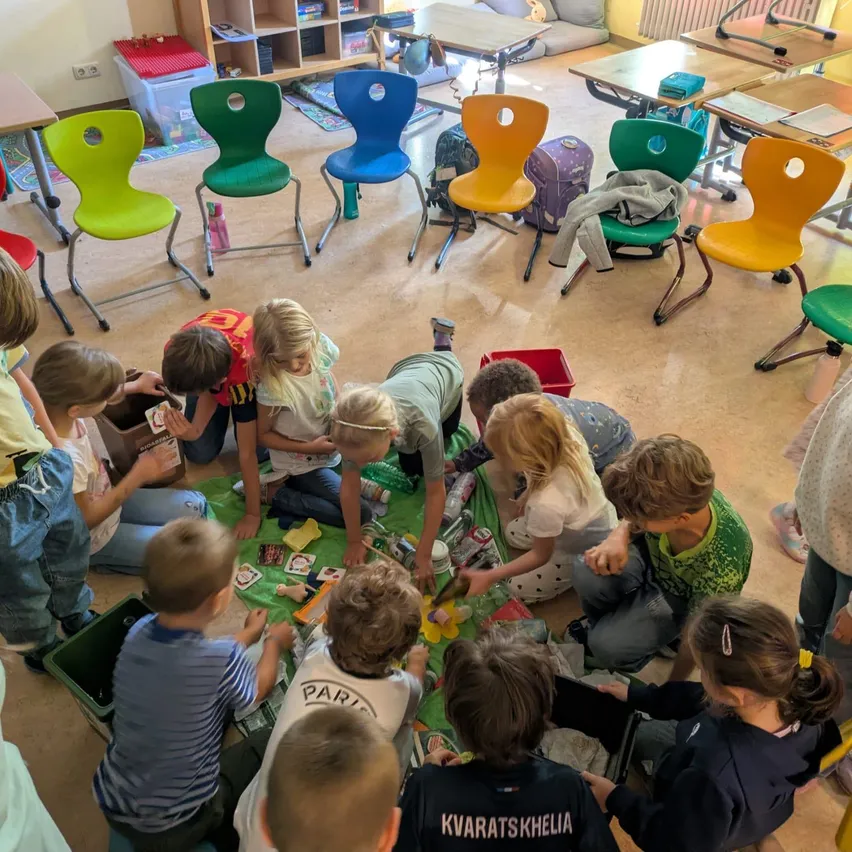 Eine Gruppe von Kindern sitzt auf dem Boden in einem Klassenzimmer und ist mit verschiedenen Spielzeugen und Karten auf einem grünen Teppich beschäftigt.