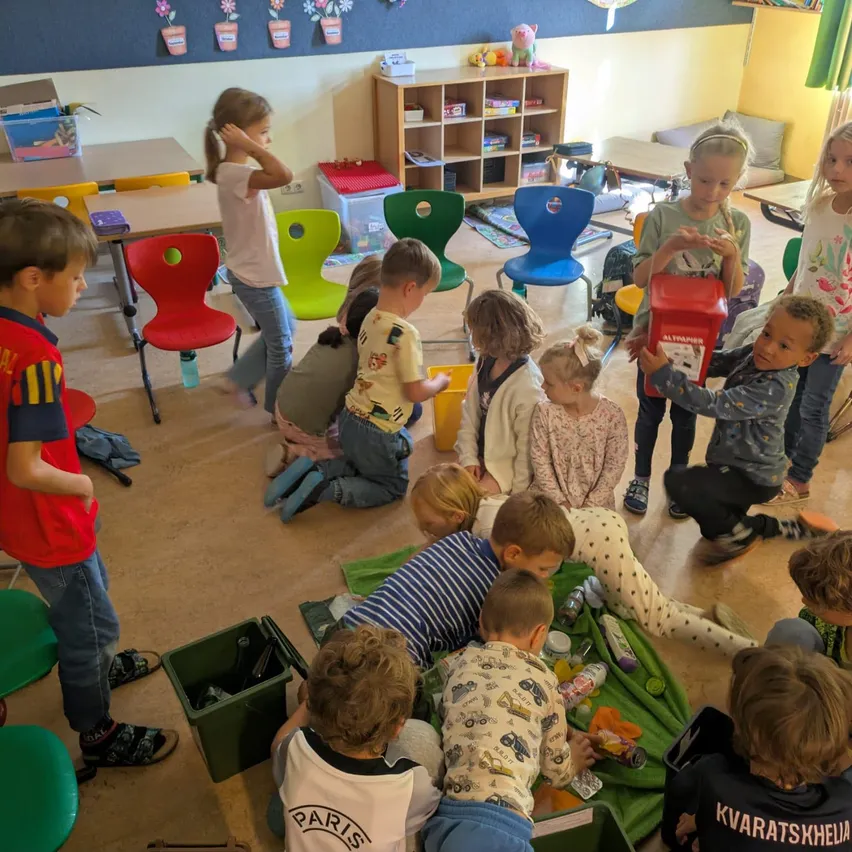 Eine Gruppe von Kindern versammelt sich in einem Klassenzimmer, einige stehen und einige sitzen auf dem Boden. Sie sind in einer Aktivität verwickelt, wobei einige rote Boxen halten. Das Klassenzimmer ist mit bunten Stühlen und Tischen gefüllt, und es gibt verschiedene Gegenstände auf den Regalen und dem Boden.