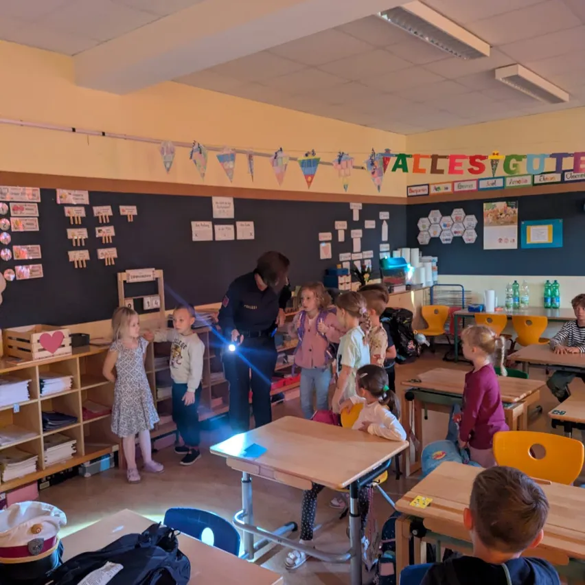 Ein Klassenzimmer mit Kindern und einem Polizeibeamten. Der Beamte hält eine Taschenlampe und spricht mit den Kindern.