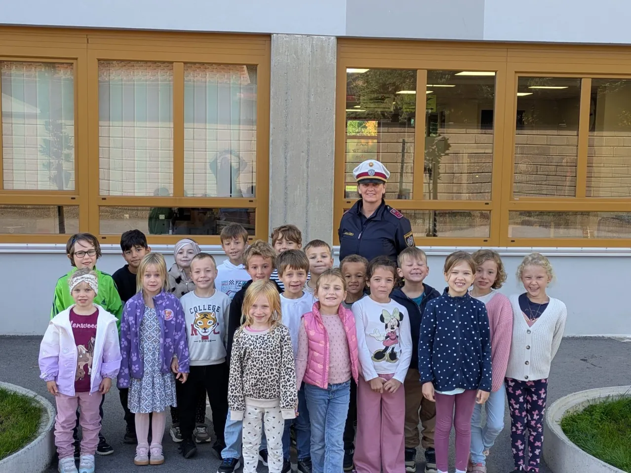 Eine Gruppe von Kindern und einer Frau in Uniform stehen vor einem Gebäude mit großen Fenstern.