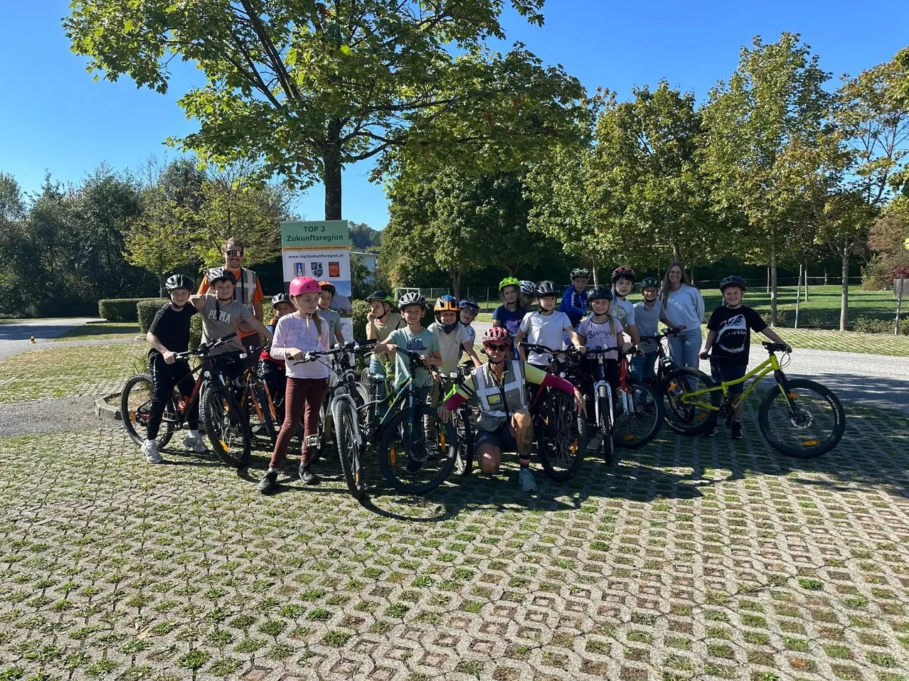 Eine Gruppe von Kindern und Erwachsenen posiert mit ihren Fahrrädern für ein Foto im Park.