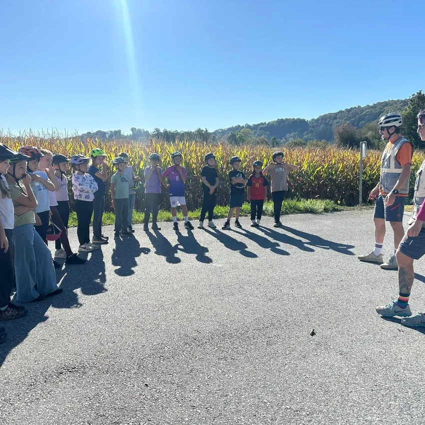 Eine Gruppe von Kindern, die Helme und Turnschuhe tragen, steht in einer Reihe auf einer Straße mit einem Maisfeld im Hintergrund.
