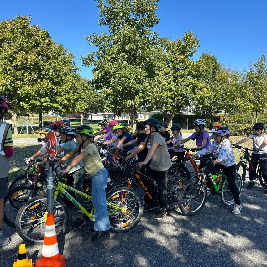 Eine Gruppe von Kindern und Erwachsenen versammelt sich auf einem Parkplatz, alle tragen Helme und fahren Fahrräder. Eine Frau in einer reflektierenden Weste steht in der Nähe, und Verkehrskegel sind auf dem Boden platziert.