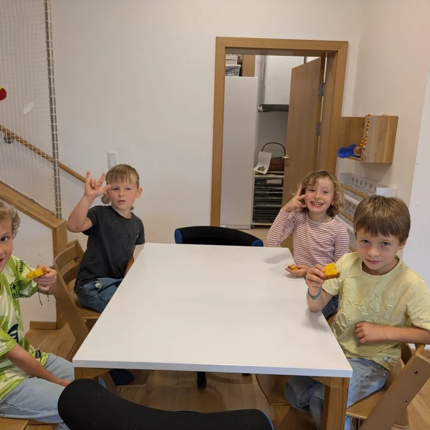 Vier junge Jungen sitzen um einen Tisch herum, lächeln und posieren für ein Foto. Sie halten Stücke von Essen in ihren Händen. Dahinter befindet sich eine Treppe, eine Tür und eine Wand mit einer Uhr und Regalen.