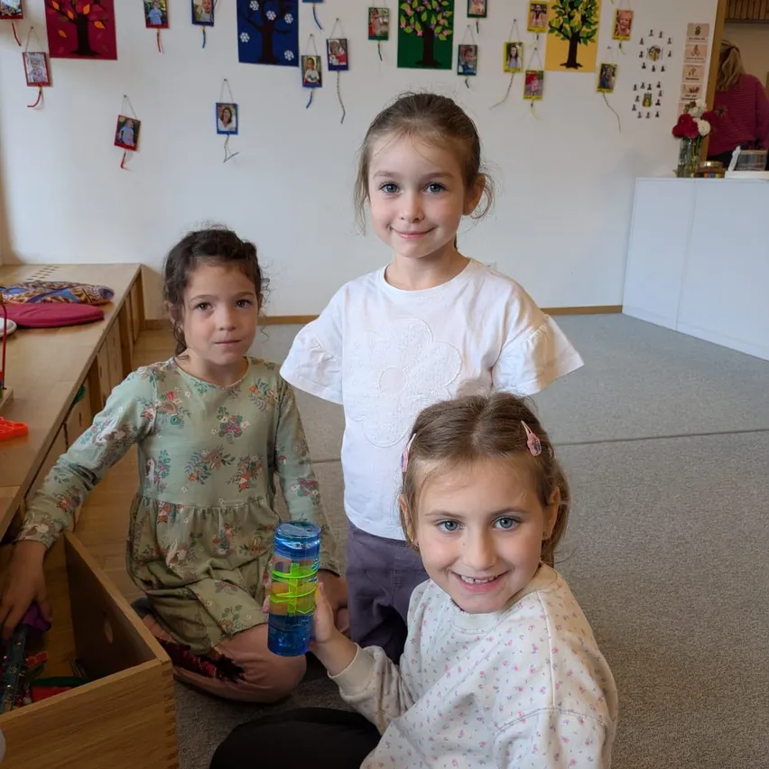 Drei junge Mädchen sitzen auf dem Boden in einem Raum. Das Mädchen auf der linken Seite hält eine blaue Flasche mit grüner Flüssigkeit darin. Das Mädchen in der Mitte lächelt und schaut in die Kamera. Das Mädchen auf der rechten Seite schaut auf die Flasche.