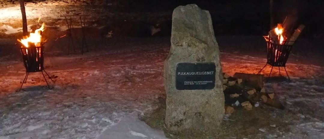 Ein Steindenkmal mit einer Plakette steht in einem verschneiten Feld, beleuchtet von zwei brennenden Lagerfeuern auf beiden Seiten. Die Lagerfeuer sind von Holzscheiten umgeben.
