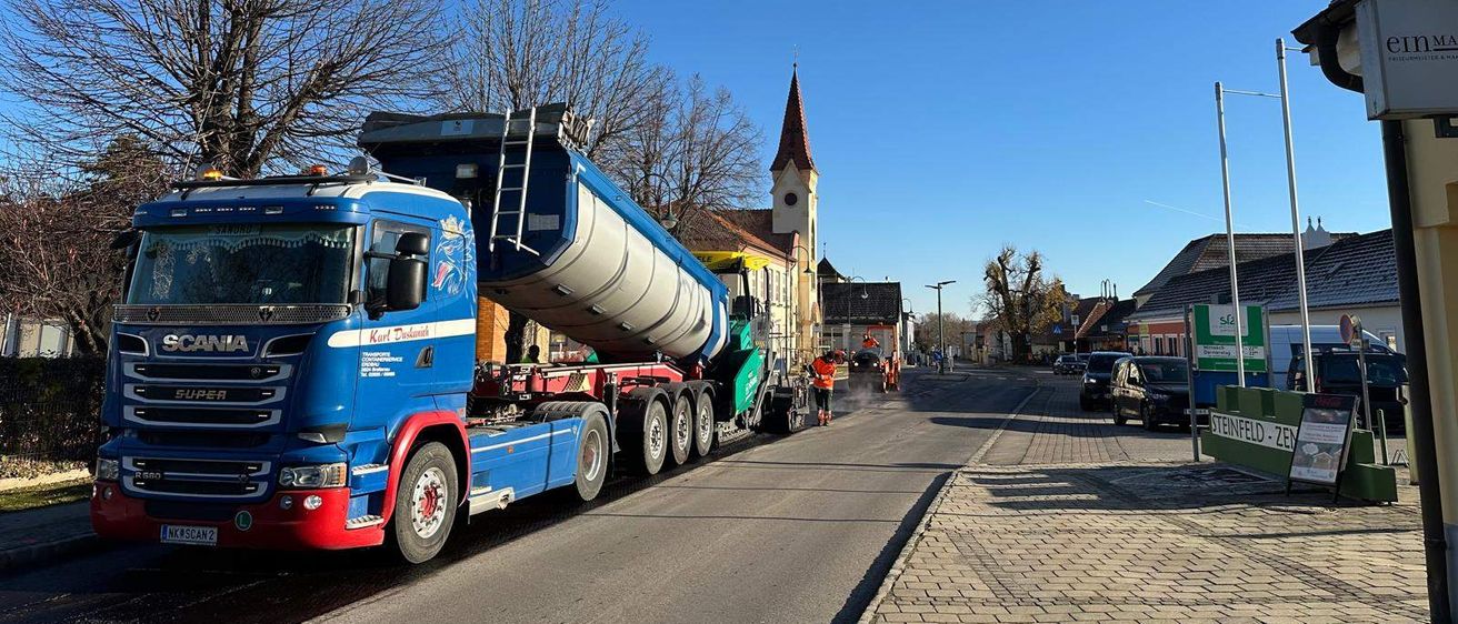 Ein großer blauer und weißer Lkw ist am Straßenrand geparkt, mit einer Person in der Nähe. Der Lkw hat einen großen Tank auf dem Anhänger. Bäume und eine Kirche sind im Hintergrund.