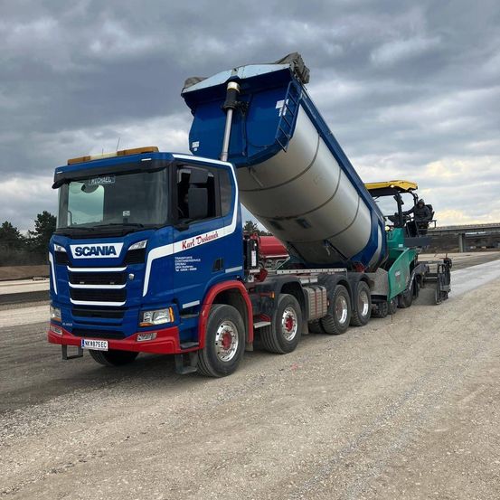 Ein blauer Scania-Lkw mit einem großen Anhänger fährt unter einem bewölkten Himmel auf einer Schotterstraße.
