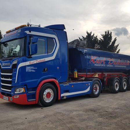 Ein blauer Karl Duskamich LKW mit einem roten Anhänger ist auf einem unbefestigten Weg mit Bäumen im Hintergrund geparkt.