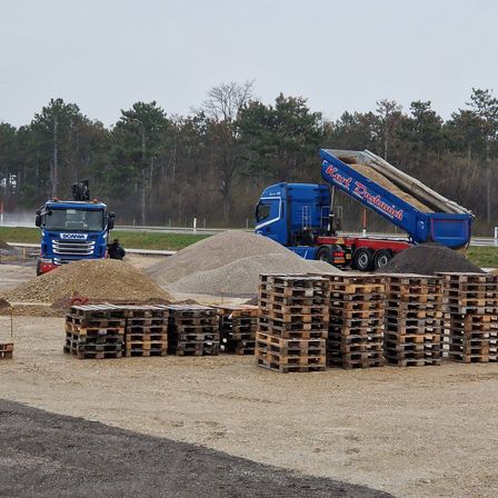 Zwei blaue Lkw, einer entlädt Kies und der andere mit einem Kran, sind auf einer Erdstraße geparkt. In der Nähe sind zahlreiche Holzpaletten gestapelt, und eine Person steht in der Nähe. Bäume und eine Straße sind im Hintergrund zu sehen.