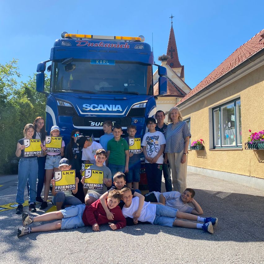 A group of children and adults pose for a photo in front of a Scania truck, holding signs that say Friends on the Road. Behind them, a building with a red roof and a church spire.