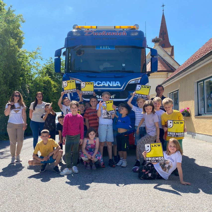 A group of children and adults stand in front of a blue truck, holding signs with the words Friends on the Road. Behind them, a church with a steeple is visible.