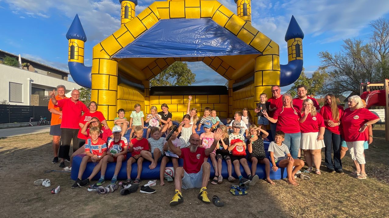 Eine Gruppe von Kindern und Erwachsenen posiert für ein Foto vor einem gelben Hüpfburg mit blauem Dach. Einige winken und lächeln.