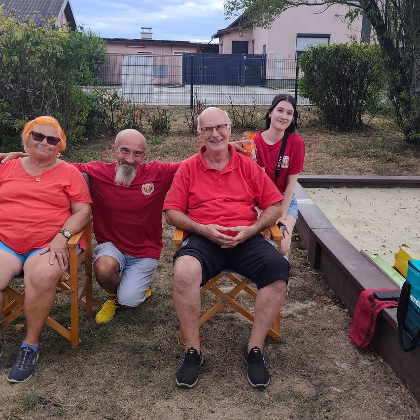 Drei Menschen in roten Shirts sitzen auf Stühlen in einem Garten, mit einem jungen Mädchen, das hinter ihnen steht.