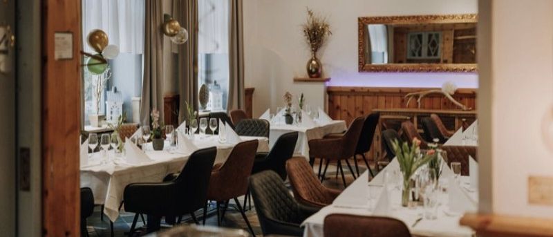 Ein Restaurant-Innenraum mit Holzwänden, einer Holzdecke und einem Spiegel an der Wand. Der Essbereich hat lange Tische mit weißen Tischdecken und einigen Weingläsern. Die Stühle sind in Reihen angeordnet, wobei einige Personen darauf sitzen.