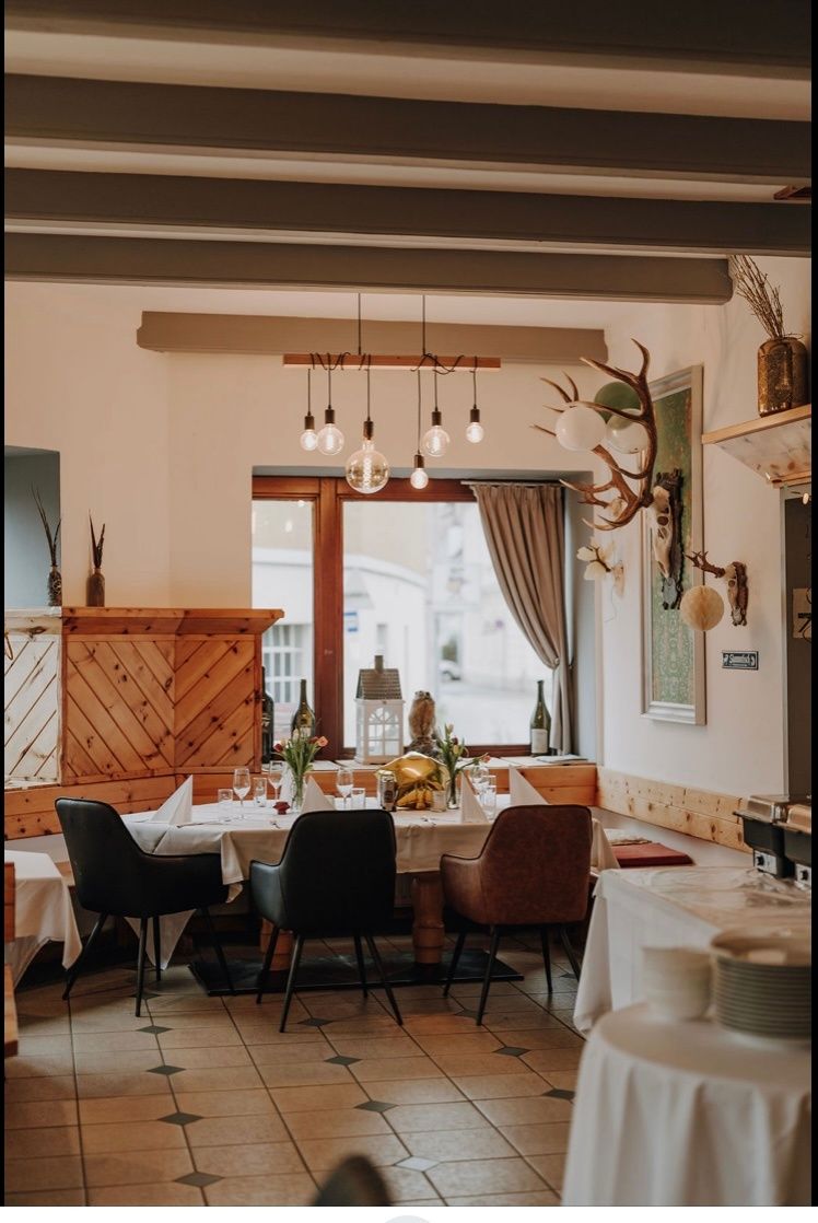 Ein Restaurant-Interieur mit Holzwänden und Decke, einem großen Fenster mit Vorhängen und einem Esstisch, der mit Blumen, Weingläsern und Tellern gedeckt ist. An der Wand befinden sich Hirschgeweihe und eine Pflanze.