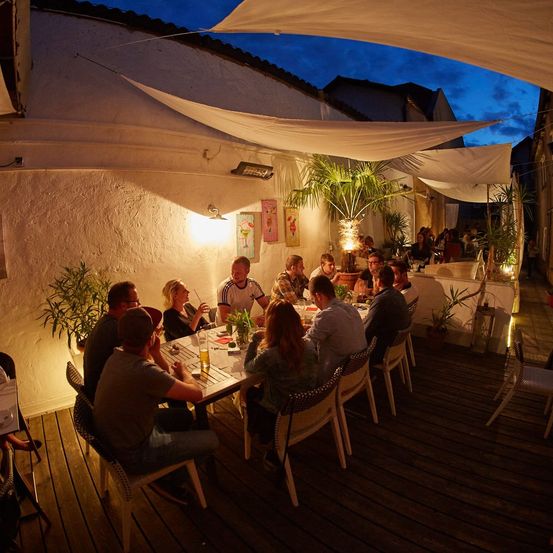 Eine Gruppe von Menschen sitzt bei Nacht auf einer Terrasse um einen Tisch herum, genießt Getränke und Gespräche. Die Terrasse ist von weißen Wänden umgeben und hat Pflanzen und Lichter.