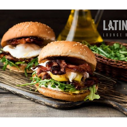 Zwei Frühstücksburger mit Ei, Speck und Grünzeug auf einem Holzschneidebrett. Eine Ölflasche und ein Korb mit Salat sind im Hintergrund. Latin Lounge-Branding.
