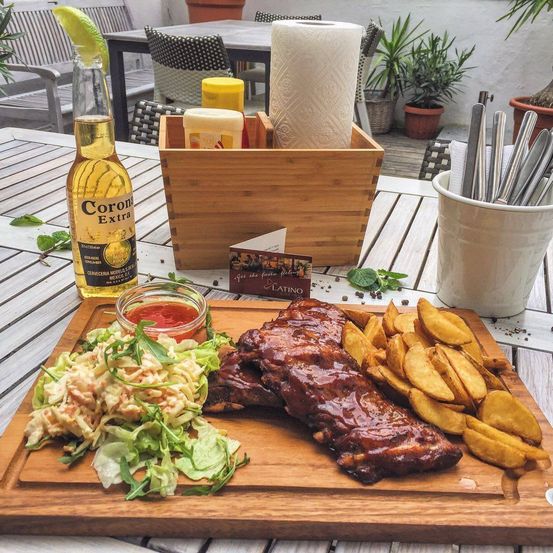 Ein Holztisch mit einem Teller mit Rippchen, Krautsalat und Pommes, neben einer Corona-Bierflasche. Eine Holzbox hält Gewürze und Papiertücher.