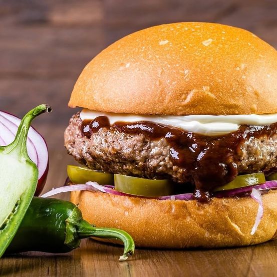 Ein Hamburger mit einem Patty, Zwiebeln, Gewürzgurken und Sauce liegt auf einer Holzoberfläche. Daneben liegen zwei grüne Paprikaschoten.