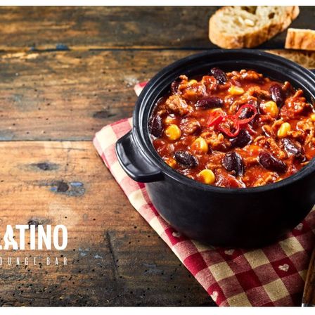 Ein schwarzer Topf mit Chili mit Bohnen und Mais auf einem karierten Tuch, auf einem Holztisch. Zwei Brotscheiben. Latin Bar.