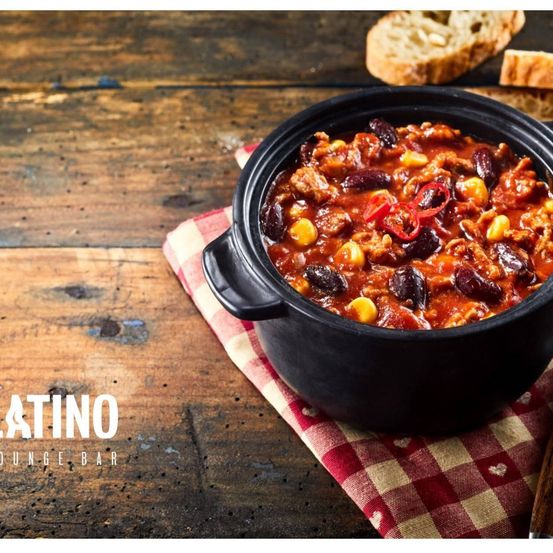 Ein schwarzer Topf mit Chili mit Bohnen und Mais auf einem karierten Tuch, auf einem Holztisch. Zwei Brotscheiben. Latin Bar.