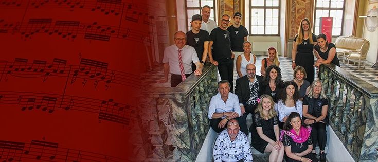Eine Gruppe von Menschen in schwarzen und weißen Outfits posiert für ein Foto auf einer Treppe. Ein roter Hintergrund hat Musiknoten.
