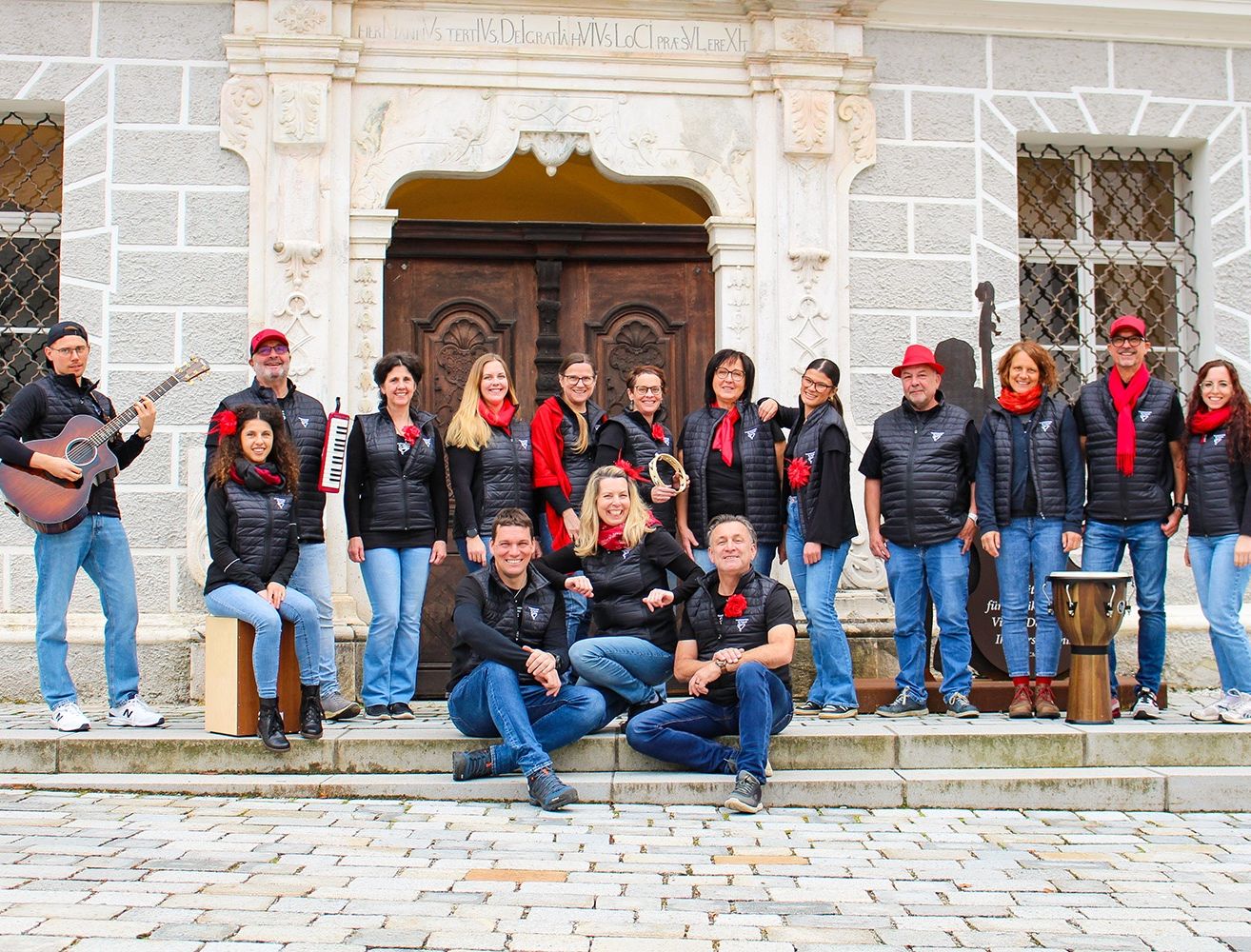 Eine Gruppe von Menschen, möglicherweise ein Chor, posiert für ein Foto vor einem Gebäude. Einige sitzen auf den Stufen, während andere hinter ihnen stehen. Sie tragen alle schwarze Jacken und rote Schals.