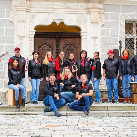 Eine Gruppe von Menschen, möglicherweise ein Chor, posiert für ein Foto vor einem Gebäude. Einige sitzen auf den Stufen, während andere hinter ihnen stehen. Sie tragen alle schwarze Jacken und rote Schals.