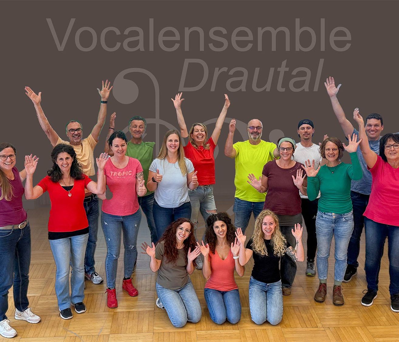 Eine Gruppe von Menschen in verschiedenen farbigen T-Shirts posiert für ein Foto auf einem Holzboden, einige stehend und andere kniend, alle lächelnd und die Hände hebend.
