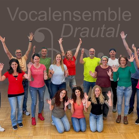 Eine Gruppe von Menschen in verschiedenen farbigen T-Shirts posiert für ein Foto auf einem Holzboden, einige stehend und andere kniend, alle lächelnd und die Hände hebend.