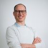 Ein lächelnder Mann mit Brille und weißem Hemd, Arme verschränkt, vor weißem Hintergrund stehend.