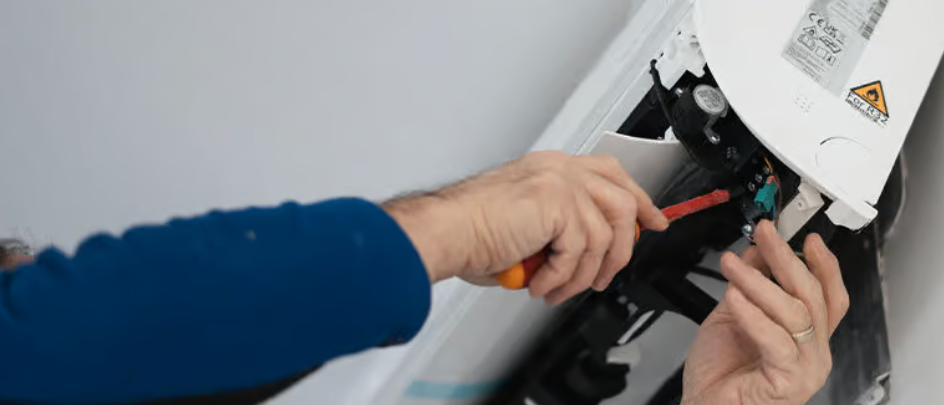 Ein Mann repariert ein Gerät an der Wand. Er hält einen Schraubendreher und scheint an der Verkabelung zu arbeiten.