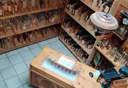 Ein Laden mit Holzregalen gefüllt mit verschiedenen Flaschen und Gläsern, einer hölzernen Theke mit Laptop, Maus und Korb und einer von der Decke hängenden Lampe.