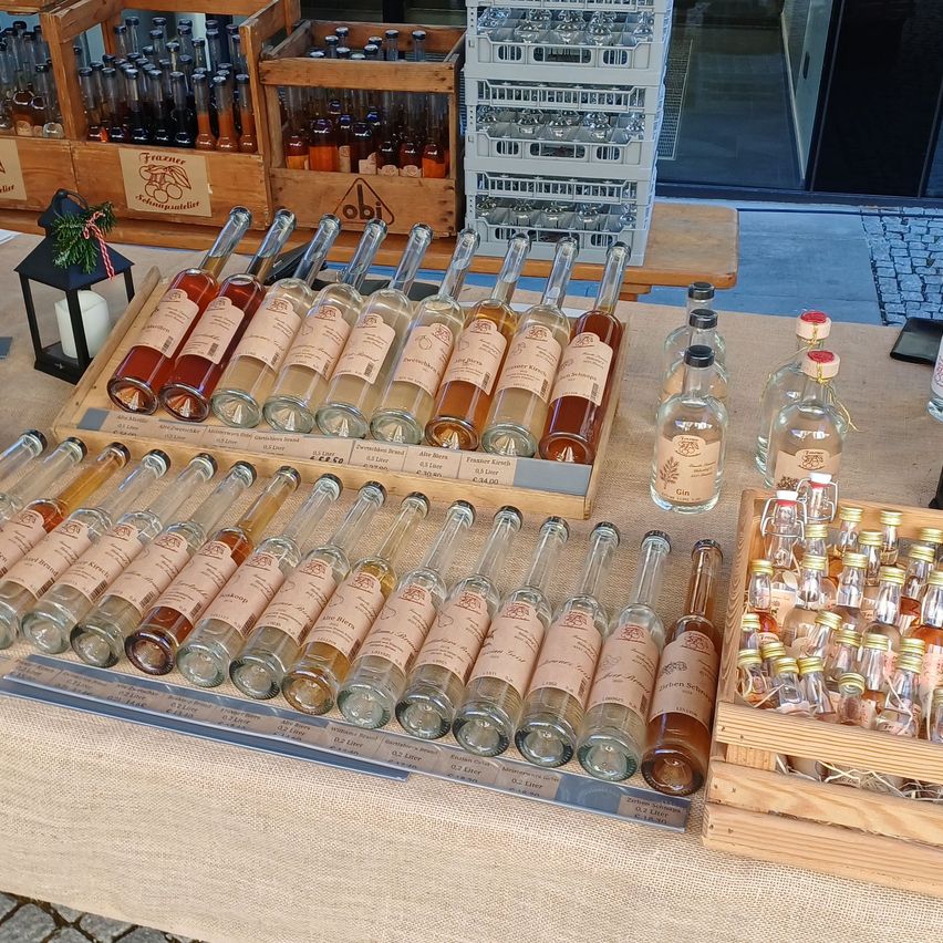 Ein Tisch zeigt eine Vielzahl kleiner Flaschen mit Schnaps und Spirituosen, die in ordentlichen Reihen angeordnet sind. Holzkisten und eine Laterne sind ebenfalls vorhanden.