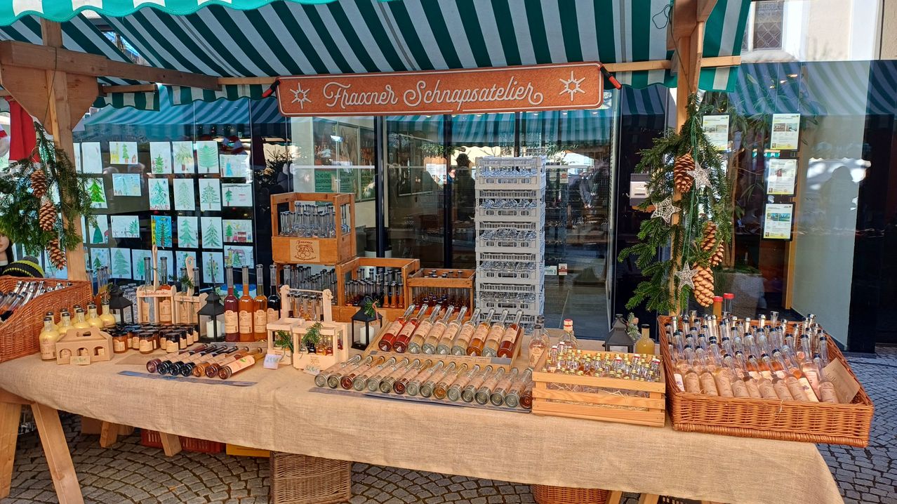 Ein Tisch mit einer Vielzahl von Spirituosen und Likören in Glasflaschen wird unter einem Baldachin mit einem Schild, das 'Fraxner Schnapsatelier' lautet, ausgestellt. Der Tisch ist mit einem Tuch bedeckt, und die Flaschen sind in Reihen angeordnet. Es gibt mehrere Aufbewahrungsboxen und Körbe mit Tannenzapfen auf dem Tisch. Der Hintergrund ist eine Glaswand mit Reflexionen von Menschen.