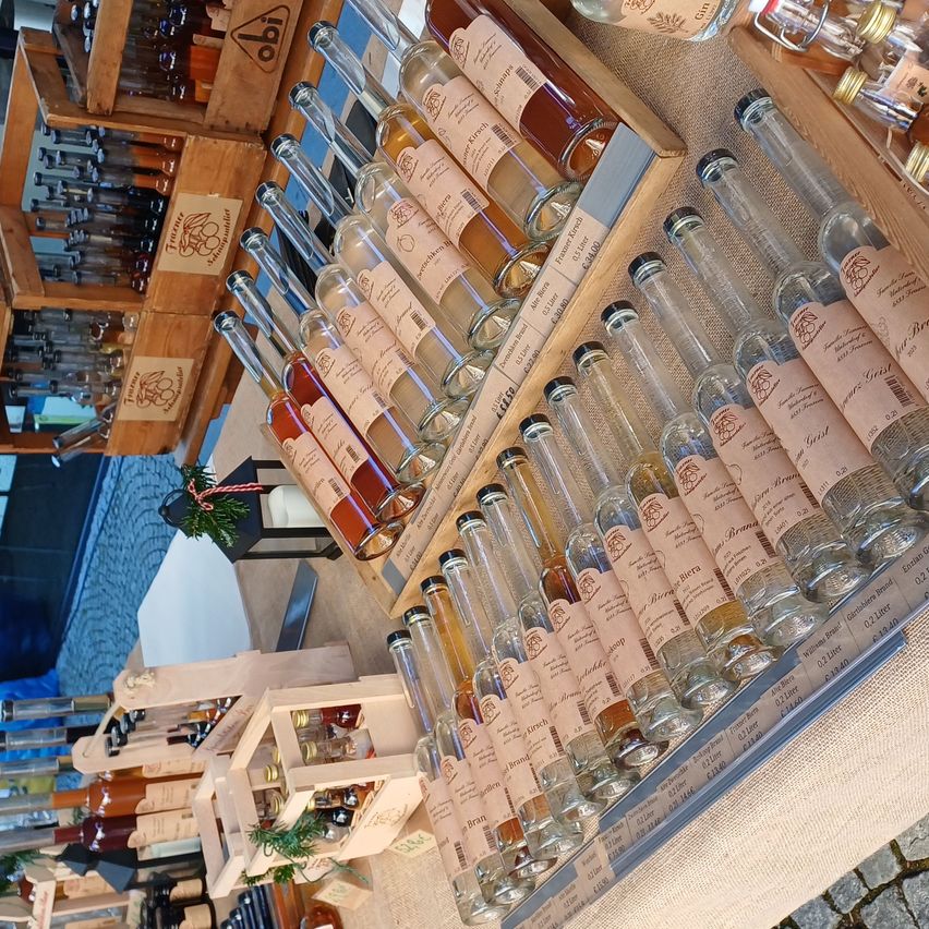 Ein Holzregal zeigt verschiedene Glasflaschen mit Etiketten, die verschiedene Arten von Alkohol in klaren, bernsteinfarbenen und dunklen Farben präsentieren. Die Flaschen sind in Reihen ordentlich angeordnet, einige auf einem Holzkasten. Im Hintergrund sind weitere Regale zu sehen, die ebenfalls mit Alkohol gefüllt sind.