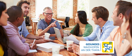 Gruppe von Menschen, die um einen Tisch mit Laptop, Tassen und Papieren sitzen. Ein Mann zeigt auf den Laptop. Das Bild hat einen Wasserzeichen mit dem Text Gesundes Waidhofen an der Thaya.